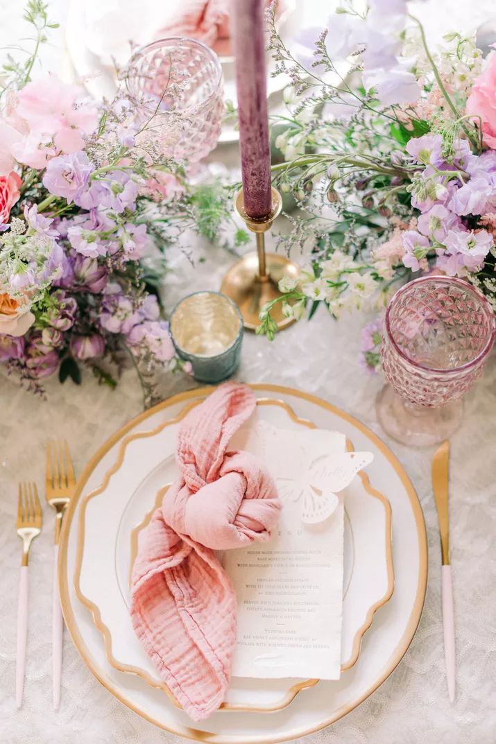 the table is set with pink and purple flowers, goldware, and napkins