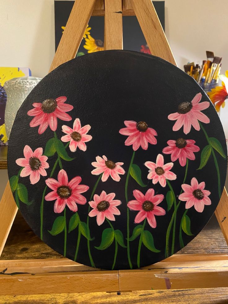 a painting of pink daisies painted on a black circle with green leaves in the center