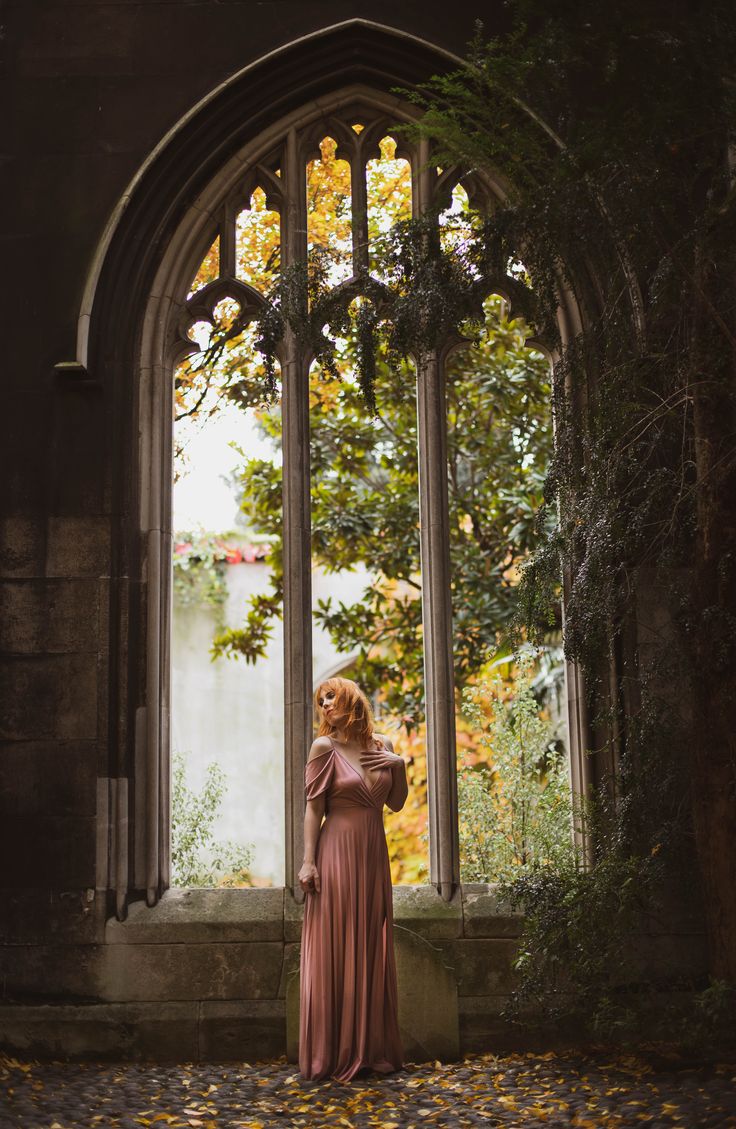 a woman standing in front of an arched window with leaves on the ground around her