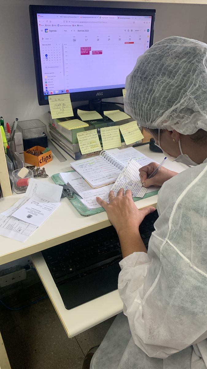 a woman in white lab coat and hat writing on paper at desk with computer monitor