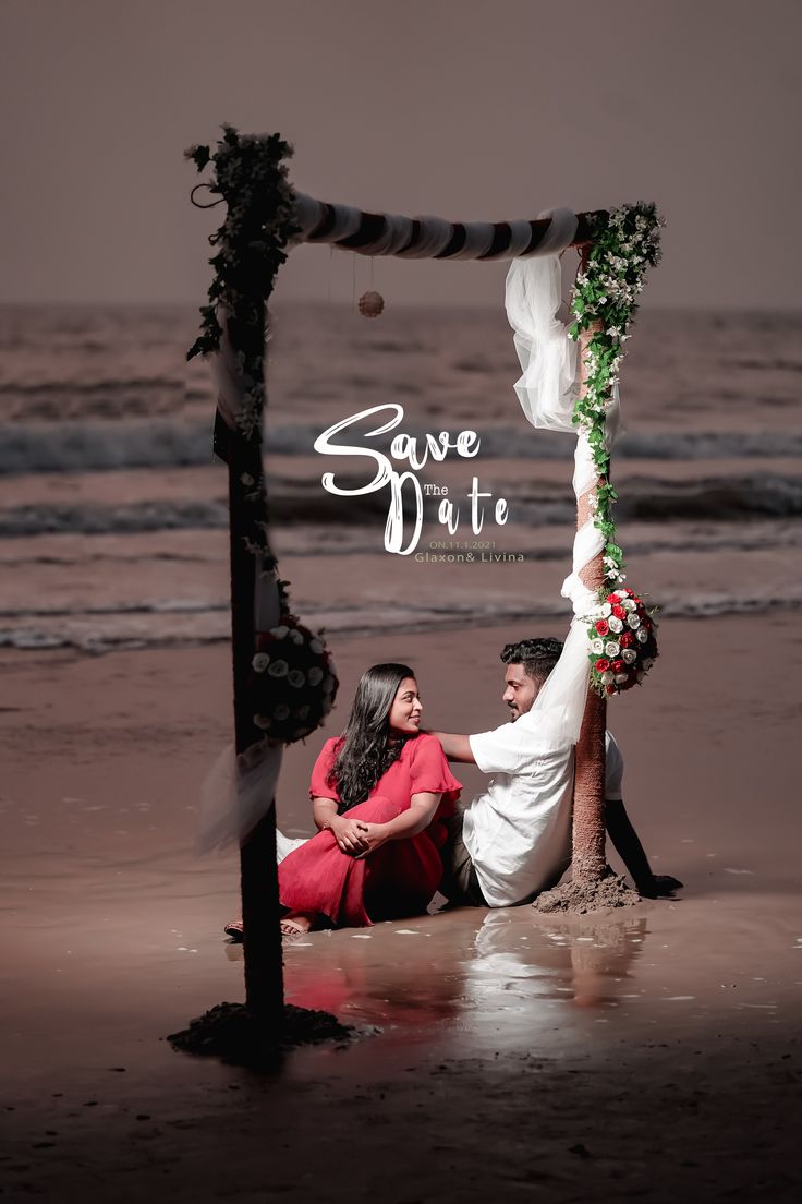 a man and woman sitting on the beach under an arch with flowers hanging from it