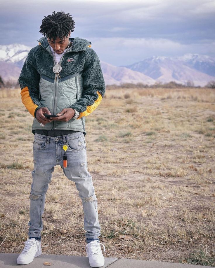 a young man standing in the middle of a field looking at his cell phone with mountains in the background