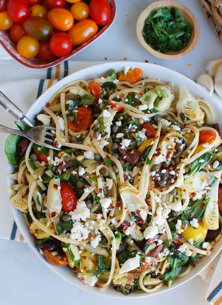 a white bowl filled with pasta and vegetables next to other bowls of tomatoes, olives, spinach, and feta cheese