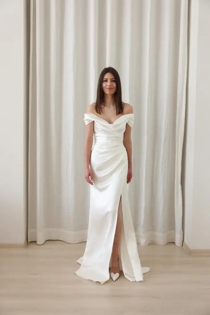 a woman standing in front of a curtain wearing a white dress with an off the shoulder slit