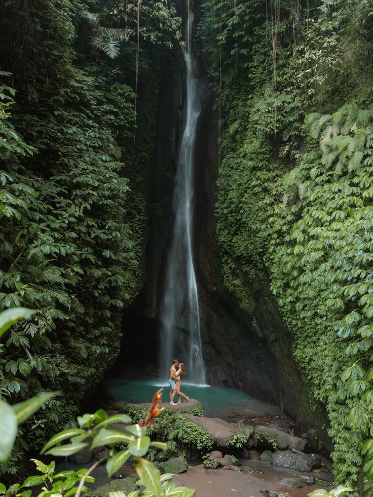 two people standing at the base of a waterfall in front of green foliage and trees