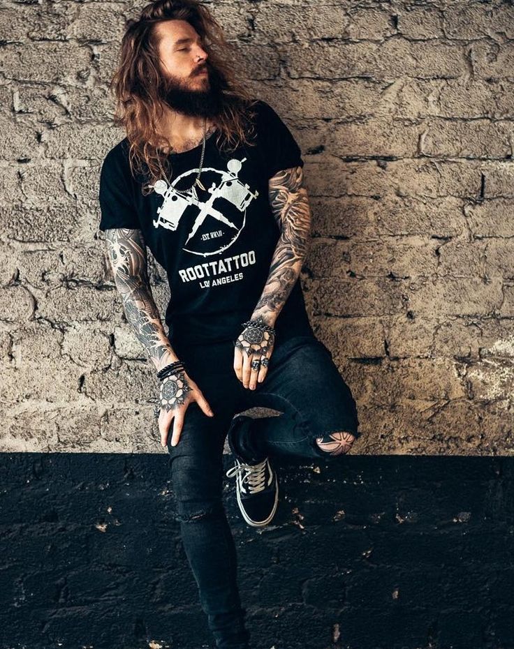 a man with long hair and tattoos sitting on a ledge next to a brick wall