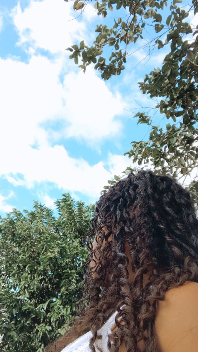 a woman with long curly hair standing in front of some trees and looking up at the sky