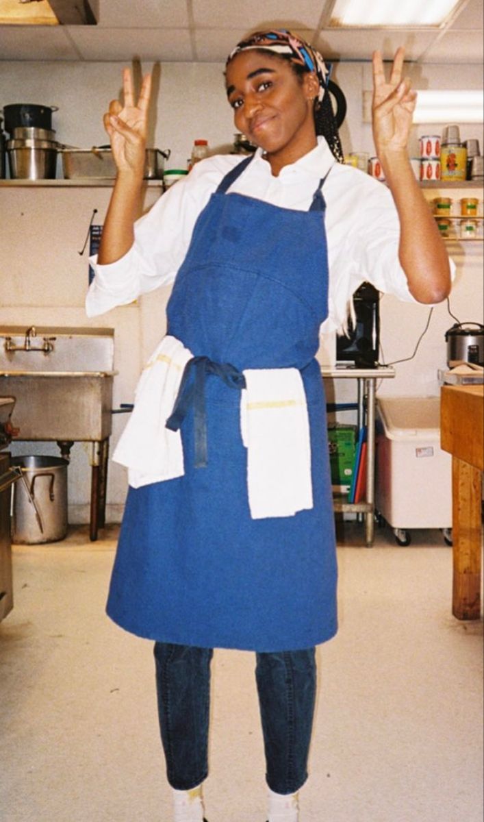 a woman standing in a kitchen with an apron on and her hands up to the side