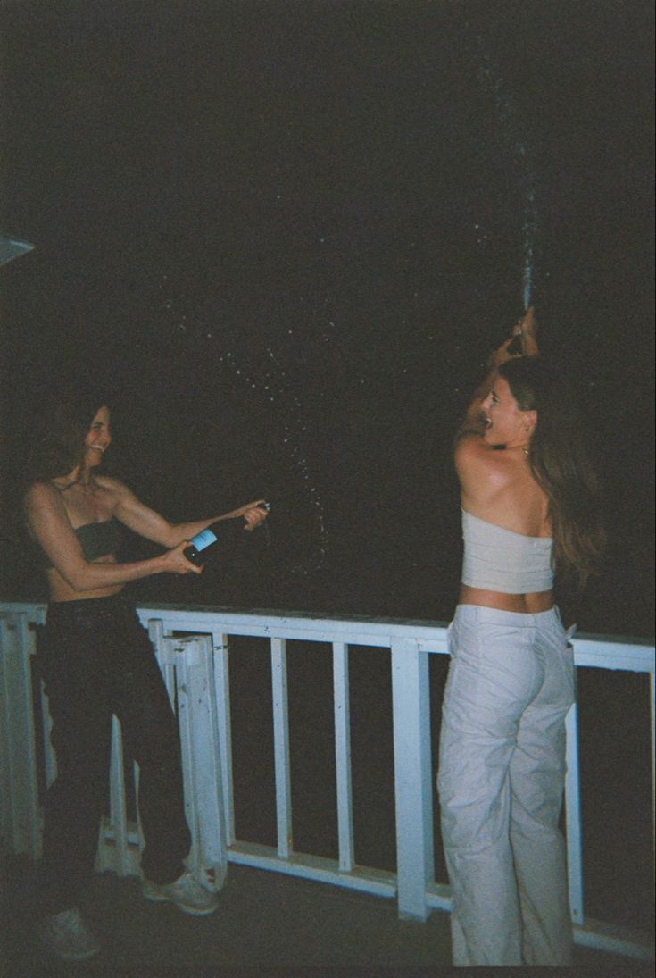 two women standing on a balcony at night, one holding a bottle and the other throwing confetti into the air