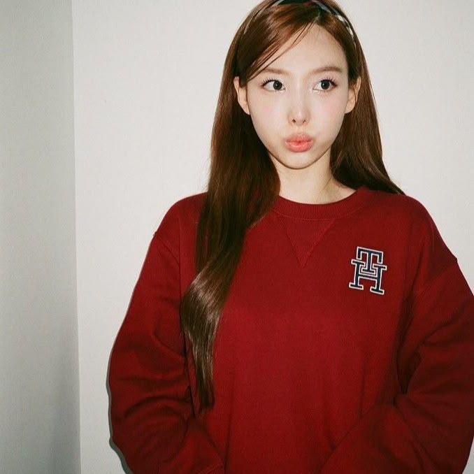 a girl with long brown hair wearing a red sweatshirt and glasses, standing in front of a white wall