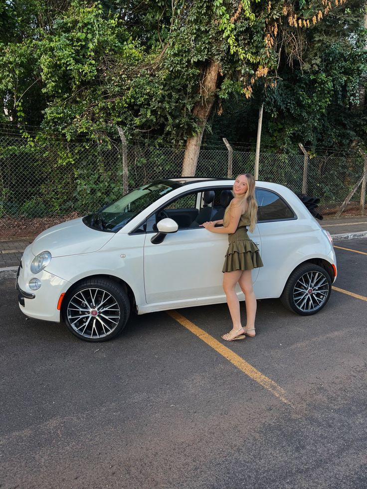 a woman standing next to a white car in a parking lot with trees behind her