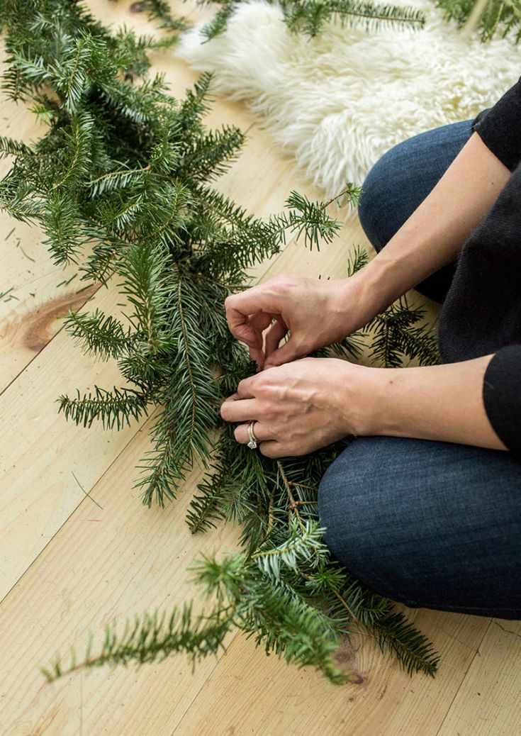 a woman is sitting on the floor making a wreath with evergreen branches and pine cones