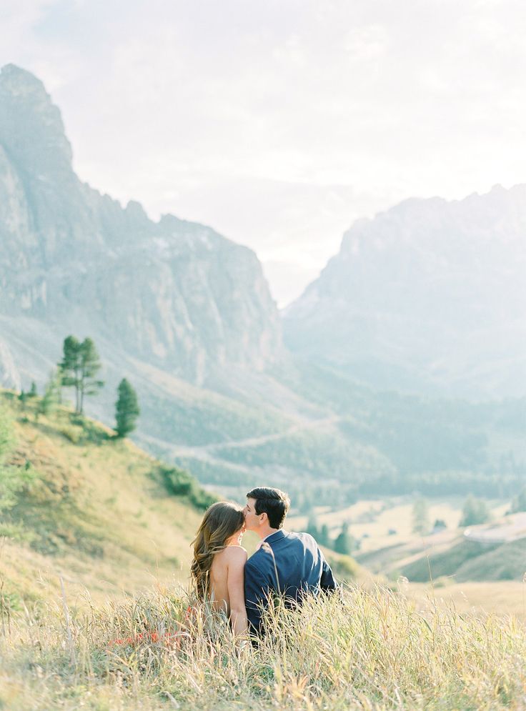 a man and woman standing in tall grass with mountains in the background