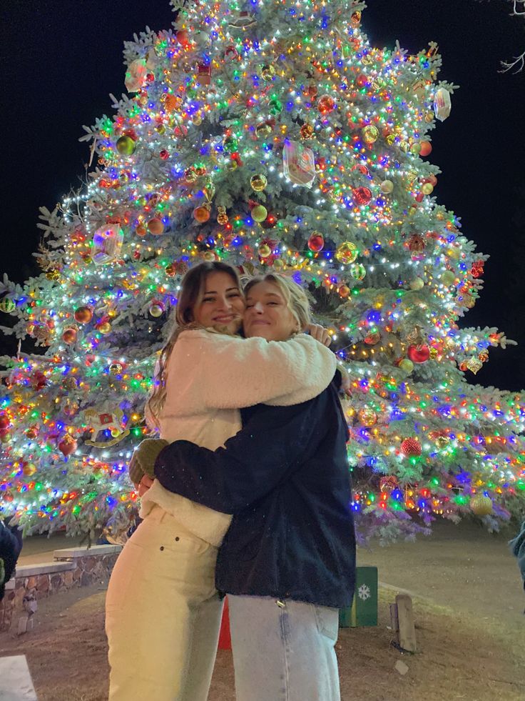 two women hugging each other in front of a christmas tree with lights all around it