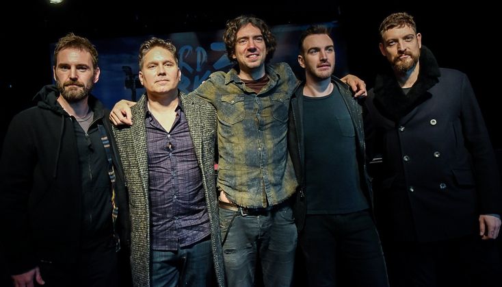 four men standing together in front of a dark background, one is wearing a jacket and the other has a sweater on