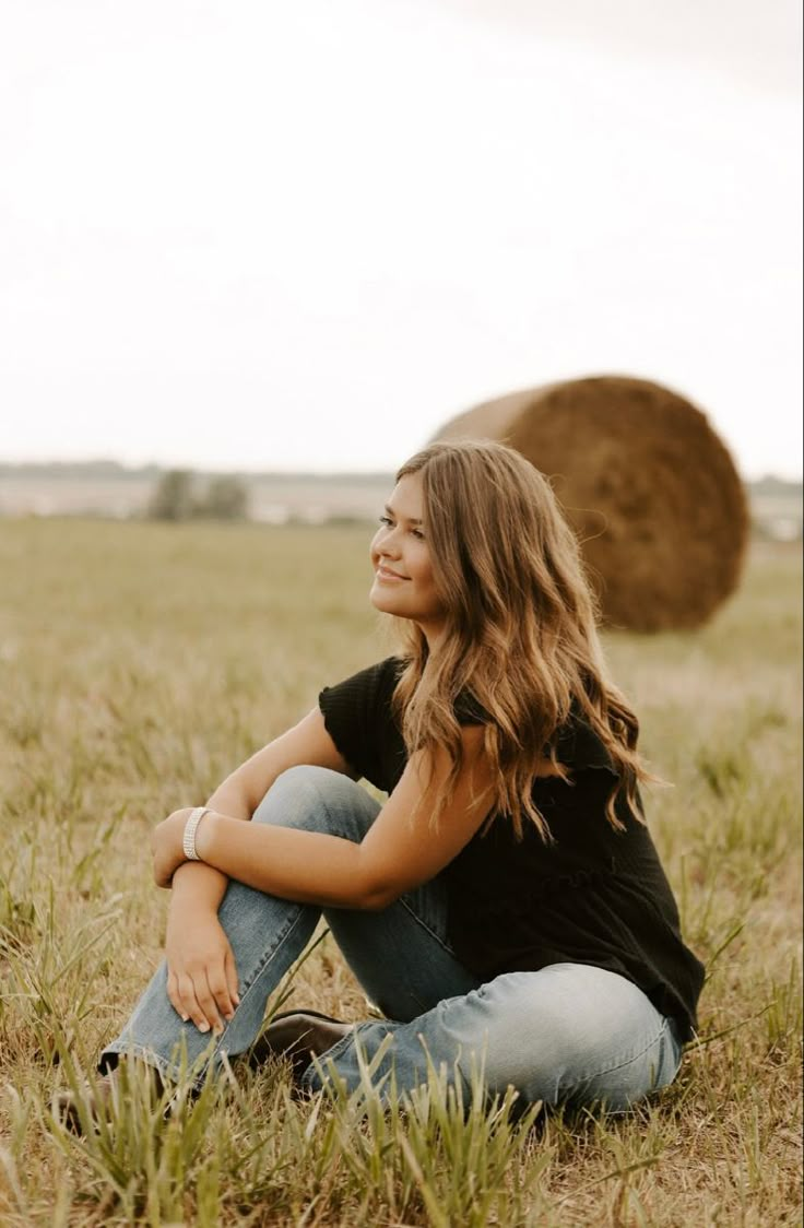 a woman sitting on the ground in a field with hay bales behind her smiling