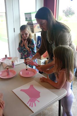 a group of children standing around a table with paper plates on it and one woman painting the plate