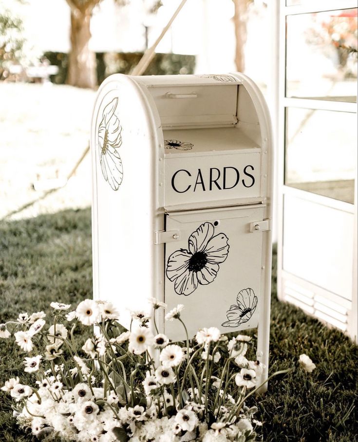 a white mailbox sitting in the grass next to some daisies and a tree