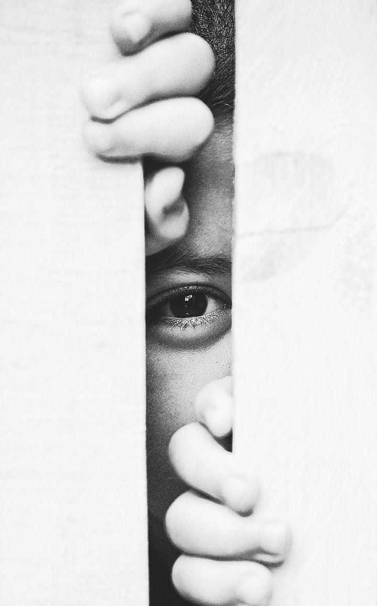 black and white photograph of a person peeking out from behind a curtain with their hands on the wall