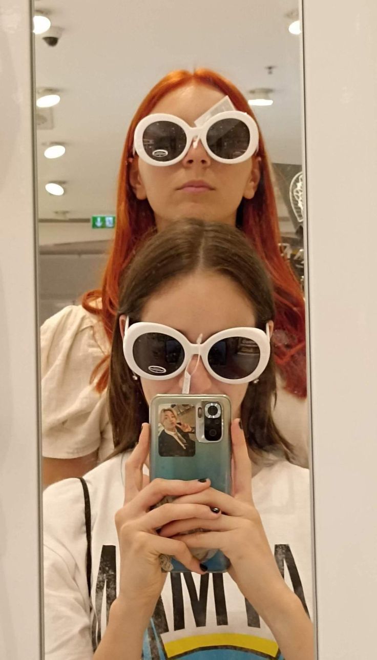 two women wearing sunglasses are looking at their cell phones in front of a mirror with the reflection of them