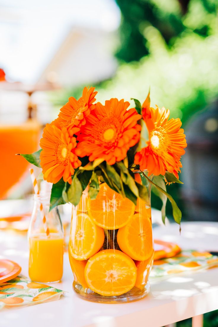 an orange flower arrangement in a glass vase on a table with plates and glasses next to it