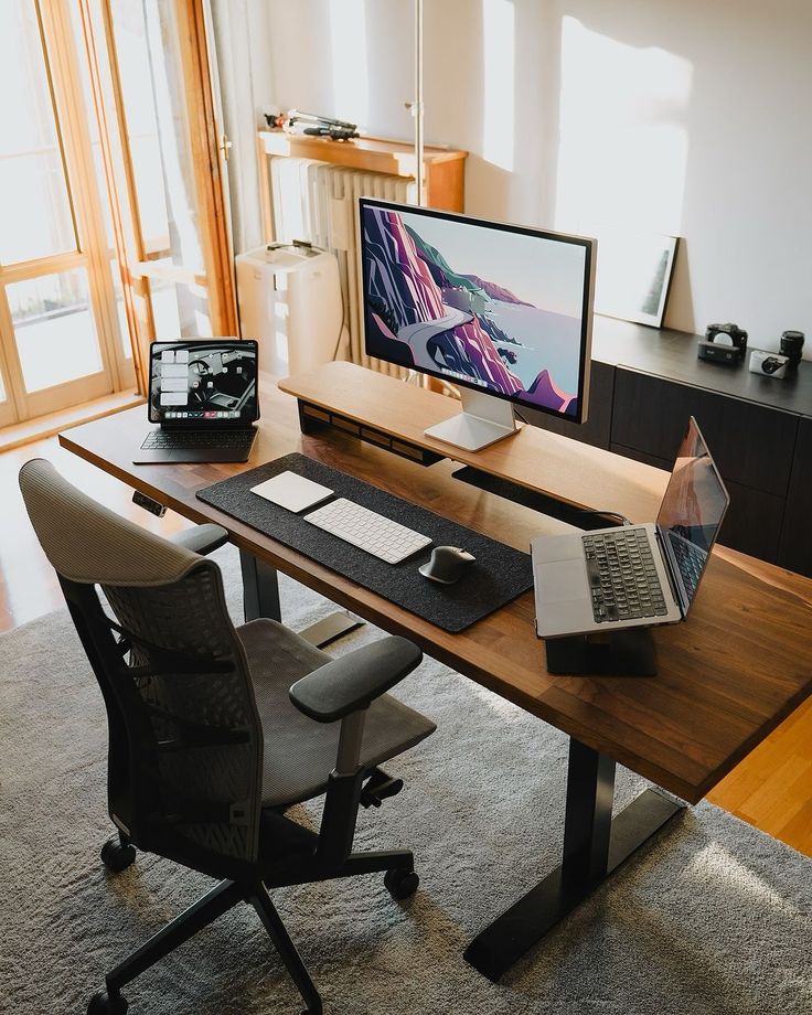 a desk with a laptop, monitor and keyboard on it in front of a window