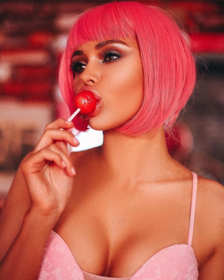 a woman with pink hair is holding a cherry and chewing on a lollipop