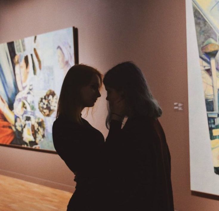 two women standing in front of paintings on the wall at an art gallery looking at each other