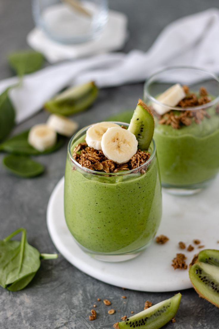 two glasses filled with green smoothie topped with banana slices and granola on a plate