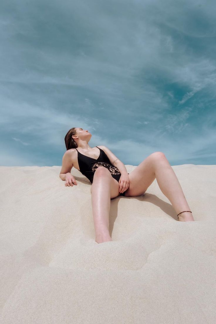 a woman in a black swimsuit laying on top of a sand dune under a blue sky