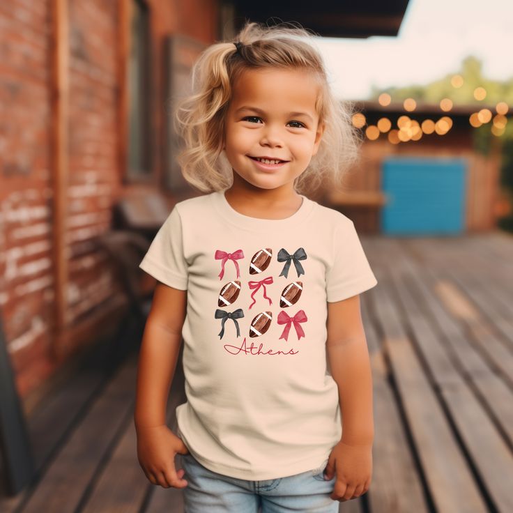 *Also available in adult sizes. See below.* Introducing our Coquette Georgia Football Toddler T-Shirt, perfect for your little ones to show their UGA team spirit! This girly tee features playful bows, sure to make your toddler stand out on game day. Go Dawgs! Cute Short Sleeve T-shirt For Game Day, Cute Cotton Game Day Top, Cute White T-shirt For Game Day, Cute Cotton Tops For Game Day, Cute Short Sleeve Tops For Playdate, Cute Cotton T-shirt For Game Day, Playful Short Sleeve T-shirt For Playdate, Cute Cotton T-shirt For Playdate, Casual T-shirt For Cheerleading