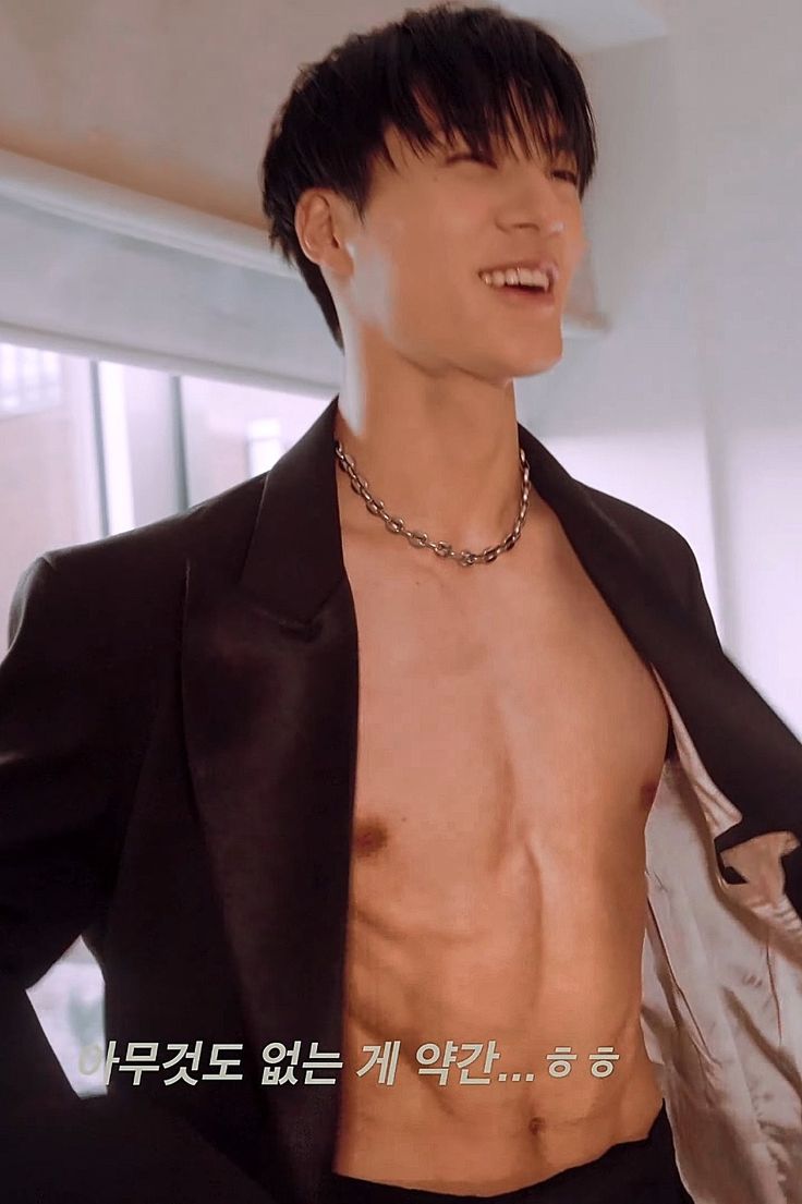 a shirtless man in a black suit and silver jacket smiles at the camera while wearing a chain around his neck