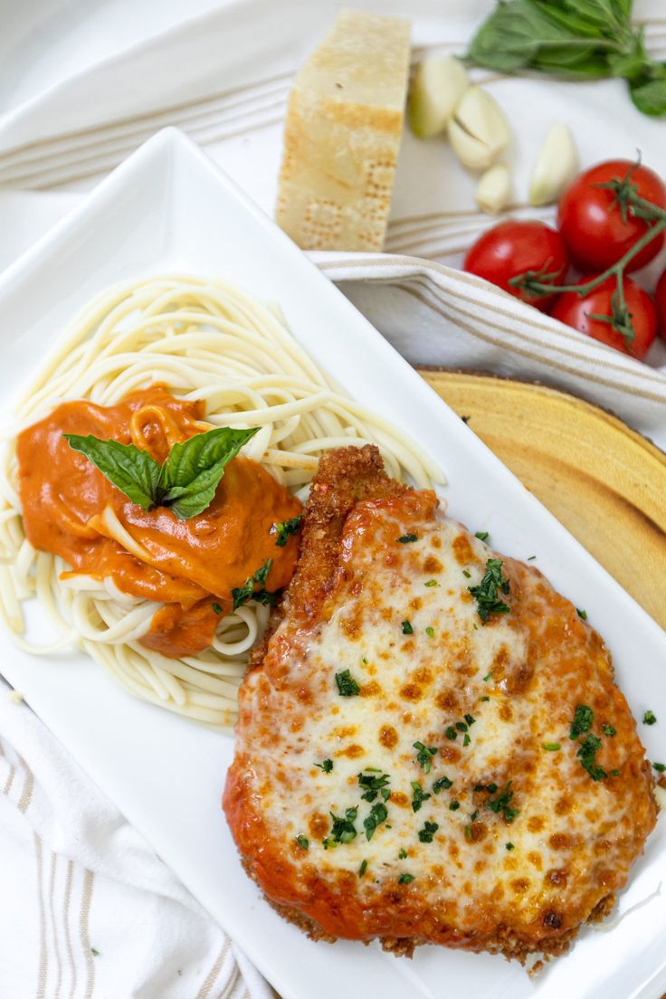 a white plate topped with meat covered in sauce and pasta next to tomatoes, bread