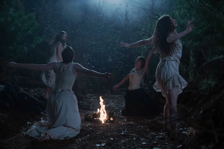 three women in white dresses are dancing around a campfire with their arms stretched out