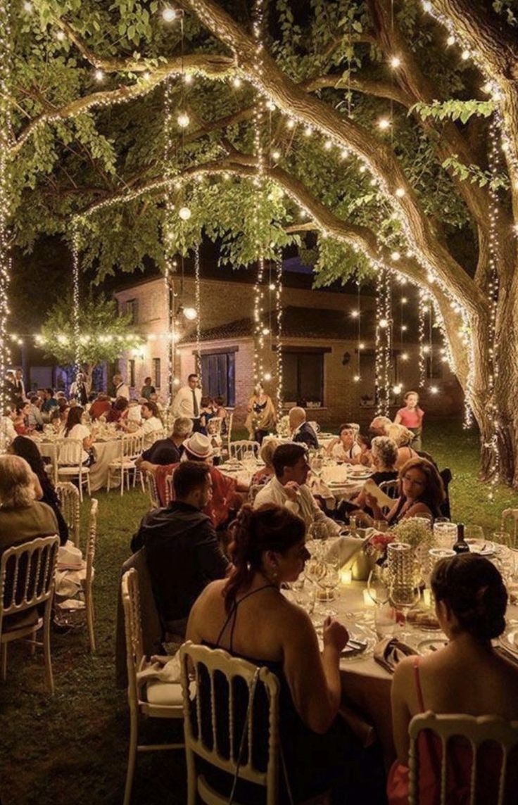 people are sitting at tables under trees with lights strung from the ceiling and on top of them