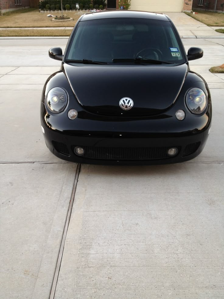 the front end of a black car parked in a driveway next to a sidewalk and house