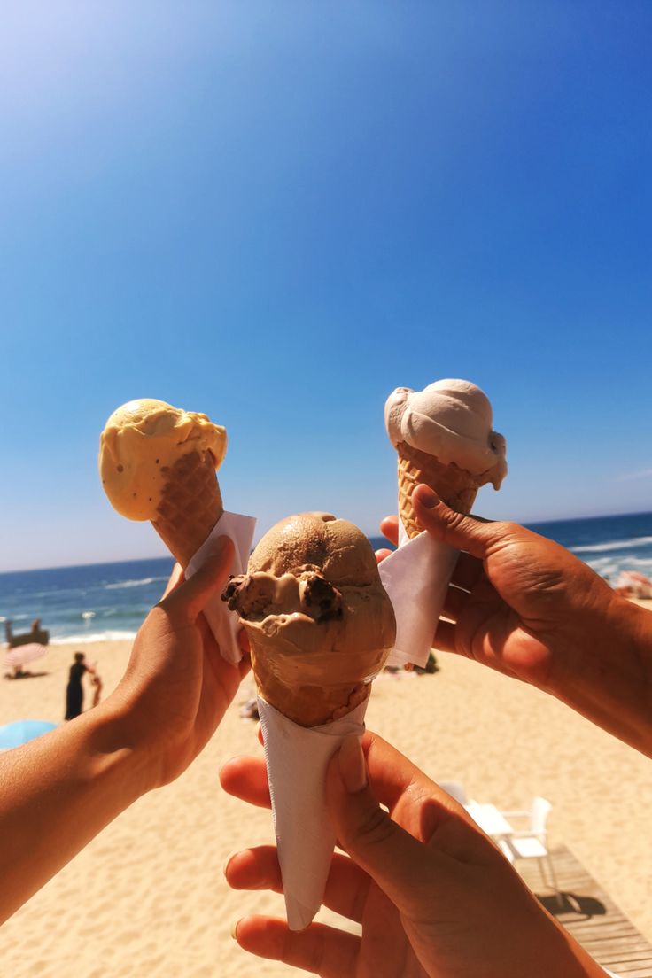 three people holding ice cream cones in their hands on the beach with ocean and blue sky behind them