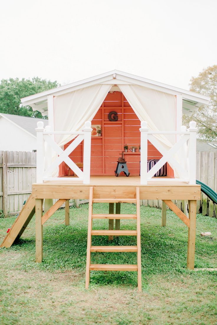 a red and white house with a ladder in the grass next to it on top of a wooden platform