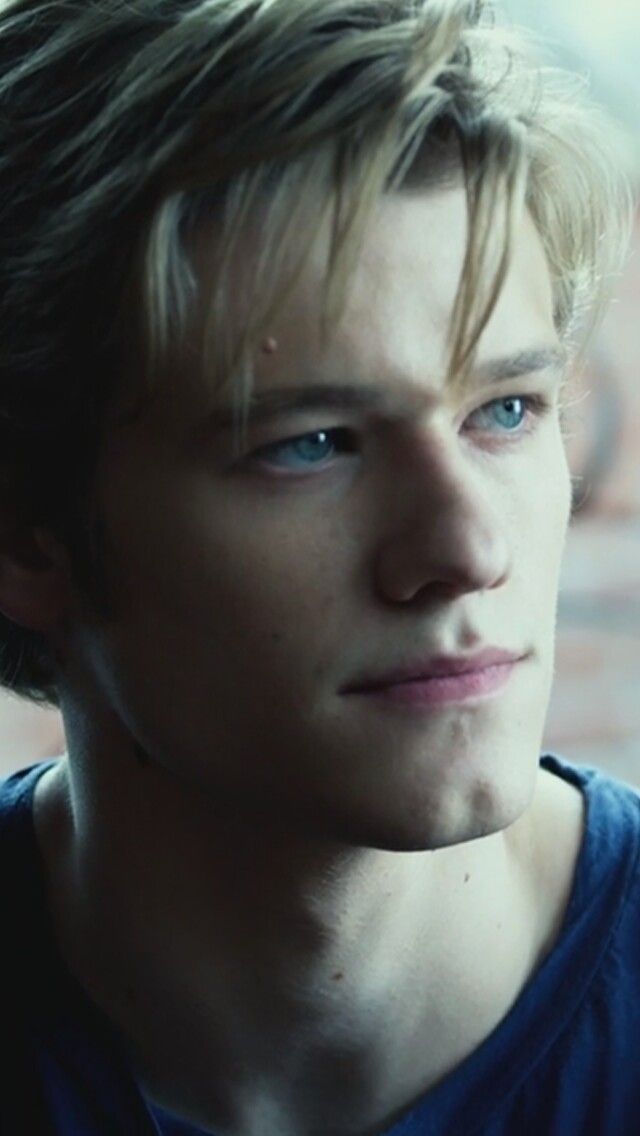 a young man with blue eyes stares into the distance while standing in front of a brick wall