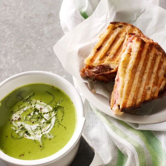 two grilled sandwiches and a bowl of green soup on a white plate with napkins