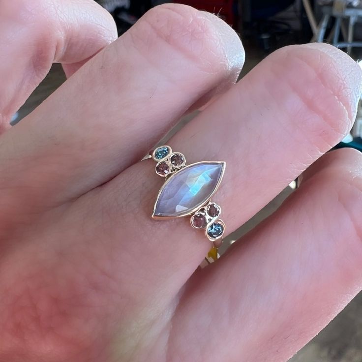 This incredible Marquis Moonstone is set between clusters of vibrant Blue Zircons and Warm Peach Sapphires. Finished with Emily's handmade Seagrass band we love it as an alternative engagement ring or right hand ring. Approximate stone size: 12mm x 6mm 2.3cts Mohs Stone Hardness: 6-6.5 This one of a kind piece is handmade to order in Emily's Hudson Valley studio. If you have questions about sizing, shipping or custom orders please reach out to us! Cluster Rose Cut Diamond Jewelry For Promise, Promise Jewelry With Cluster Of Rose Cut Diamonds, Yellow Gold Multi-stone Moonstone Jewelry, Multi-stone Sapphire Rings Marquise Cut, Marquise Cut Sapphire Multi-stone Rings, Oval Multi-stone Moonstone Rings, Heirloom Oval Moonstone Ring With Multi-stone, Unique Moonstone Ring With Rose Cut Diamonds For Anniversary, Oval Moonstone Multi-stone Rings