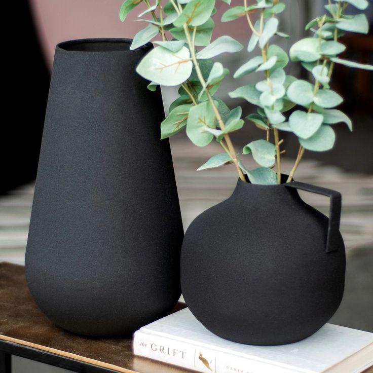 two black vases sitting on top of a table next to a book and plant