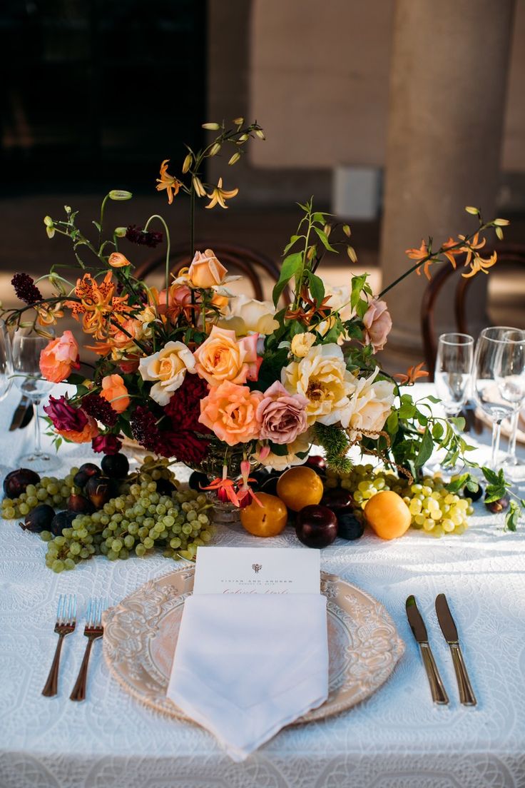 an arrangement of flowers, fruit and greenery is displayed on a table with place settings