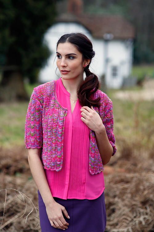 a woman standing in a field wearing a pink top and purple pants with her hand on her hip