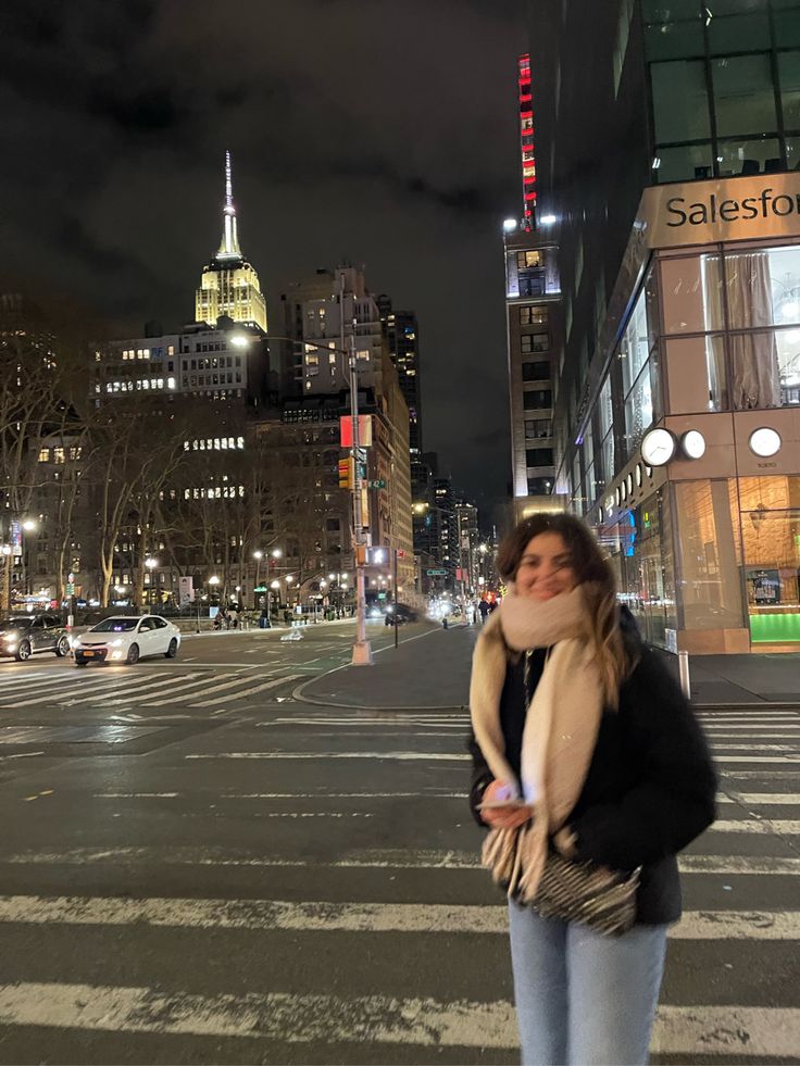 a woman standing in the middle of a crosswalk at night with buildings and lights behind her