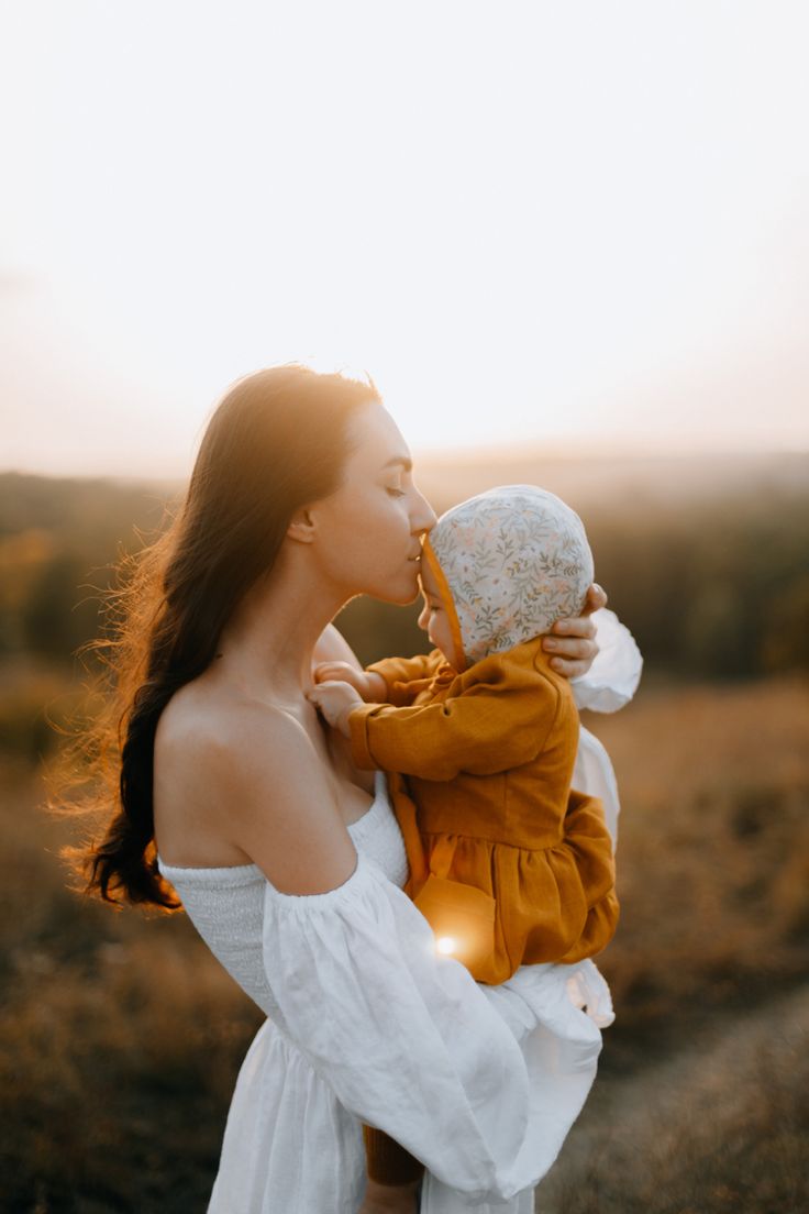 a woman holding a baby in her arms and kissing it's face with the sun behind her