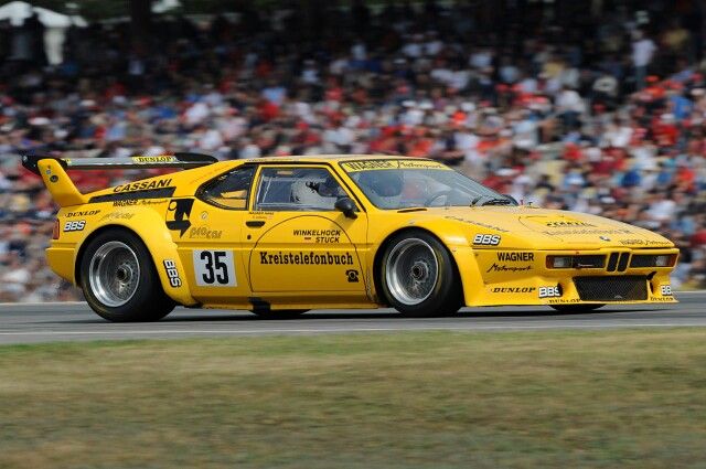 a yellow sports car driving down a race track with spectators in the stands behind it