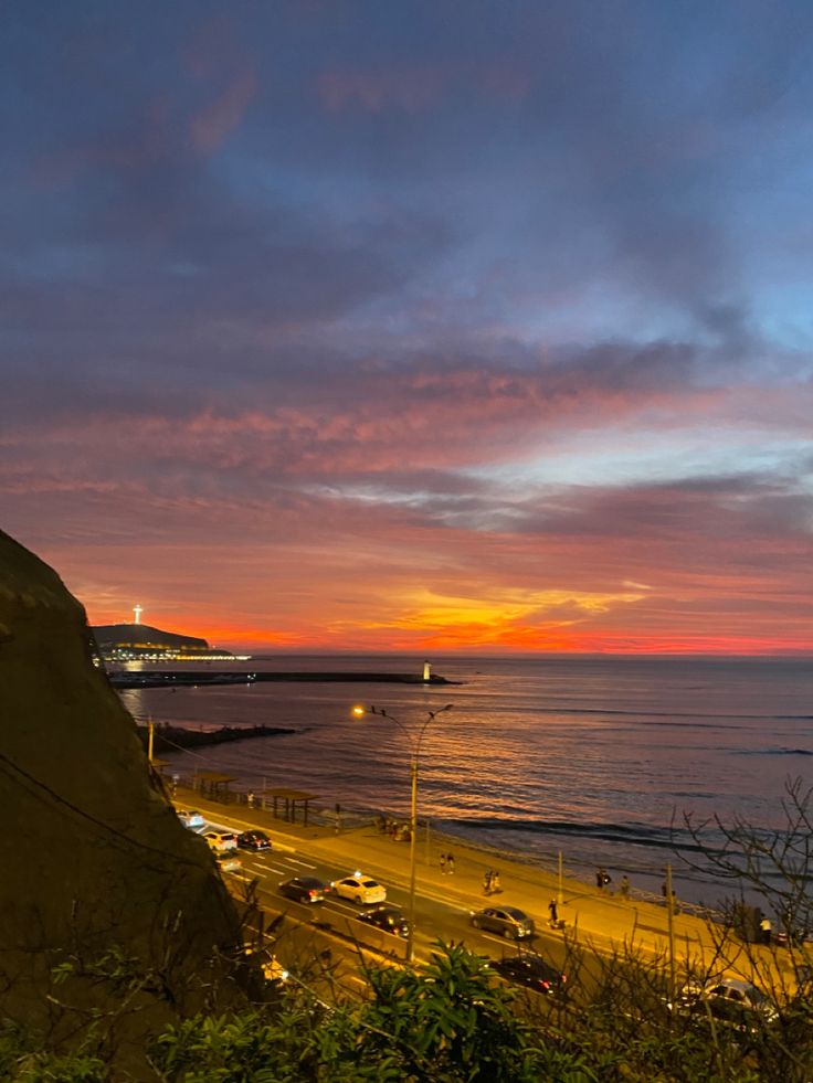 the sun is setting over the ocean and cars are parked on the beach below it