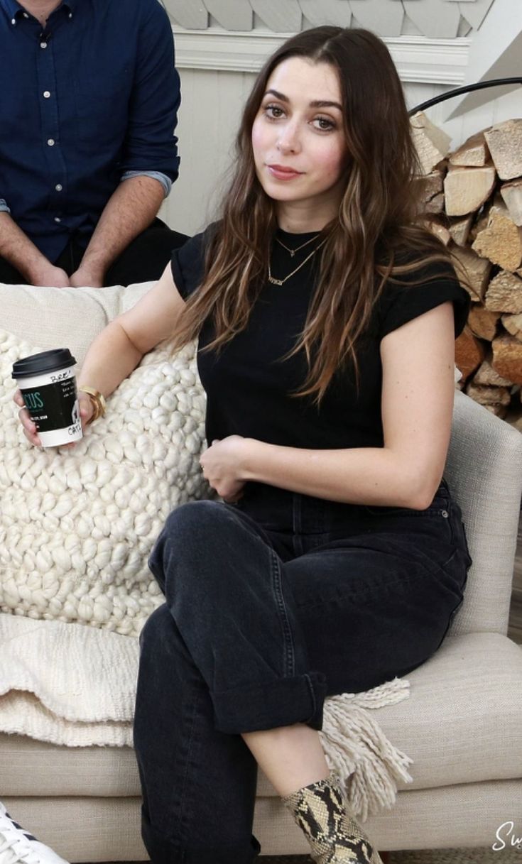 a woman sitting on top of a couch next to a pile of firewood with a cup of coffee in her hand