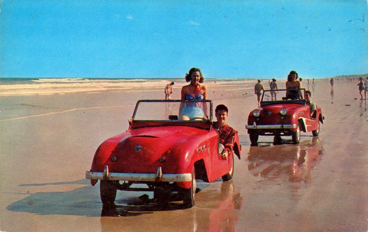 two children are riding in an old red convertible car on the beach with their parents